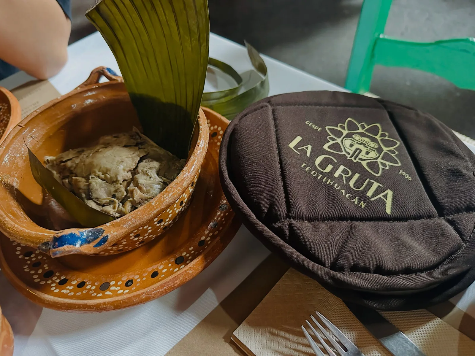 Mixiote de barbacoa servido en cazuela de barro sobre hoja de maguey, junto al tortillero bordado con el logotipo de La Gruta Teotihuacán, desde 1906